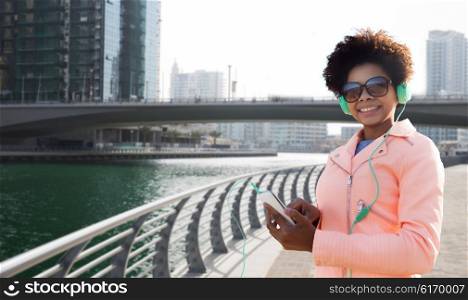 technology, lifestyle and people concept - smiling african american young woman with smartphone and headphones listening to music over dubai city street or waterfront and bridge background