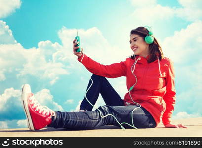 technology, lifestyle and people concept - smiling young woman or teenage girl with smartphone and headphones listening to music outdoors over blue sky and clouds background