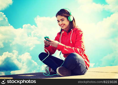 technology, lifestyle and people concept - smiling young woman or teenage girl with smartphone and headphones listening to music over blue sky and clouds background