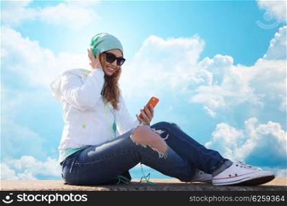 technology, lifestyle and people concept - smiling young woman or teenage girl with smartphone and headphones listening to music over blue sky and clouds background