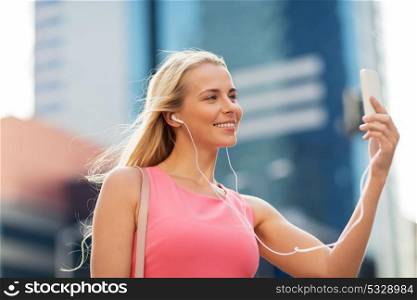technology, lifestyle and people concept - smiling young woman with smartphone and earphones listening to music and taking selfie in city. happy young woman with smartphone and earphones