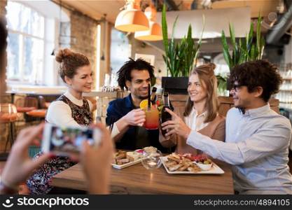 technology, lifestyle, holidays and people concept - happy friends clinking drinks and photographing by smartphone at bar or cafe