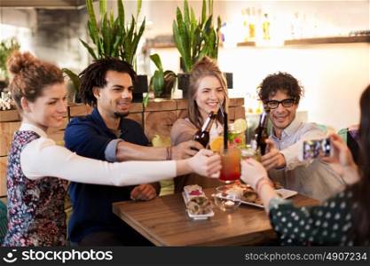 technology, lifestyle, holidays and people concept - happy friends clinking drinks and photographing by smartphone at bar or cafe. happy friends clinking drinks at bar or cafe