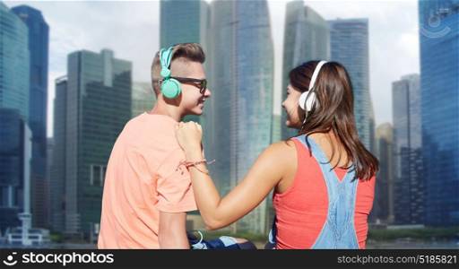 technology, love and people concept - happy teenage couple with headphones listening to music over singapore city skyscrapers background. teenage couple with headphones on river berth