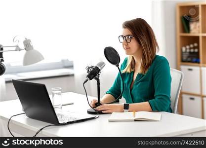 technology, mass media and people concept - woman in glasses with microphone and laptop computer talking and recording podcast at studio. woman with microphone recording podcast at studio
