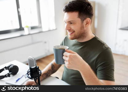 technology, mass media and podcast concept - happy male audio blogger with microphone drinking coffee and broadcasting at home office. blogger with coffee and microphone audio blogging