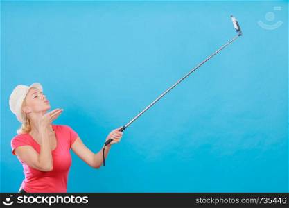 Technology, modern photography, confidence conept. Happy attractive adult blonde woman with sun hat taking picture of herself with smartphone on selfie stick.. Woman taking picture of herself with phone on stick