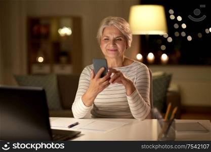 technology, old age and communication concept - happy senior woman with smartphone and papers at home in evening. senior woman with smartphone at home at night