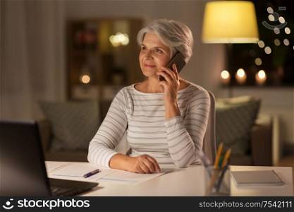 technology, old age and communication concept - senior woman with laptop computer and papers calling on smartphone at home in evening. old woman calling on smartphone at home at night