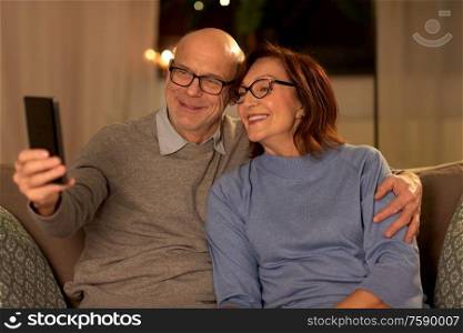 technology, old age and people concept - happy senior couple taking selfie with smartphone at home in evening. old couple taking selfie with smartphone at home