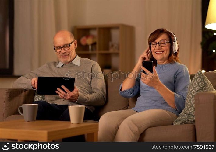 technology, old age and people concept - happy senior couple with tablet pc computer, headphones and smartphone at home in evening. happy senior couple with gadgets at home