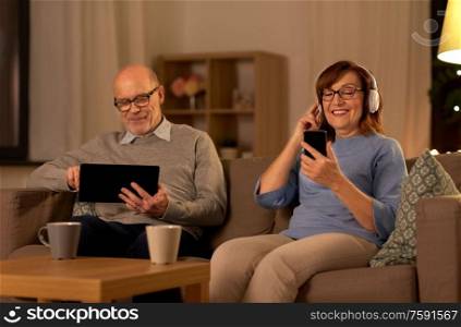 technology, old age and people concept - happy senior couple with tablet pc computer, headphones and smartphone at home in evening. happy senior couple with gadgets at home