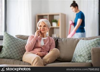 technology, old age and people concept - happy senior woman calling on smartphone and housekeeper cleaning at home. happy senior woman calling on smartphone at home
