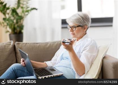 technology, old age and people concept - happy senior woman in glasses with laptop computer and smartphone using voice command recorder at home. old woman with laptop records voice by smartphone