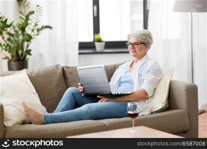 technology, old age and people concept - happy senior woman in glasses with laptop computer and glass of wine at home. senior woman with on laptop resting at home