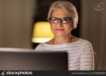 technology, old age and people concept - happy senior woman in glasses with laptop at home in evening. happy senior woman with laptop at home in evening