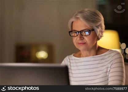 technology, old age and people concept - happy senior woman in glasses with laptop at home in evening. happy senior woman with laptop at home in evening
