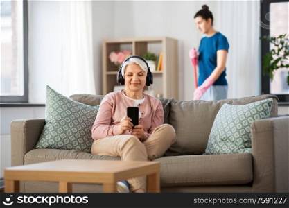 technology, old age and people concept - happy senior woman in headphones with smartphone and housekeeper cleaning at home. old woman in headphones with smartphone at home