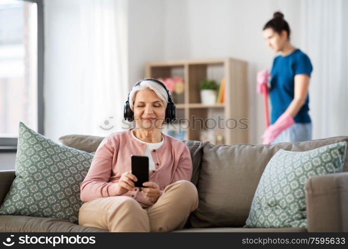 technology, old age and people concept - happy senior woman in headphones with smartphone and housekeeper cleaning at home. old woman in headphones with smartphone at home