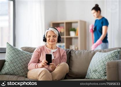 technology, old age and people concept - happy senior woman in headphones with smartphone and housekeeper cleaning at home. old woman in headphones with smartphone at home