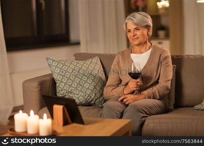 technology, old age and people concept - happy senior woman watching movie on tablet pc computer and drinking red wine at home in evening. senior woman with tablet pc drinking wine at home