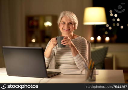 technology, old age and people concept - happy senior woman with laptop drinking coffee at home in evening. senior woman with laptop drinking coffee at home