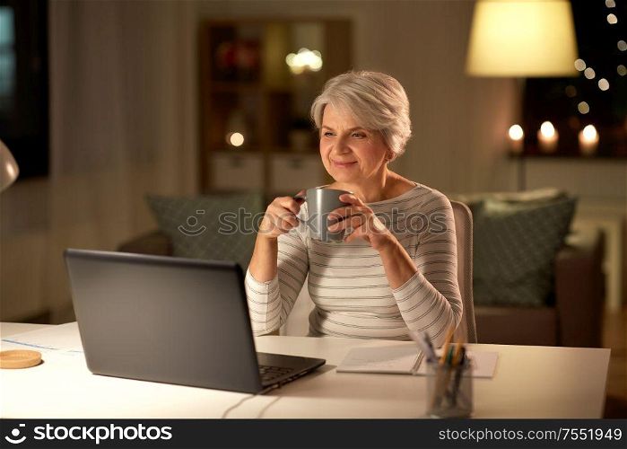 technology, old age and people concept - happy senior woman with laptop drinking coffee at home in evening. senior woman with laptop drinking coffee at home
