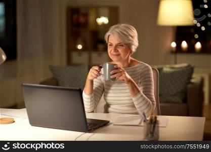 technology, old age and people concept - happy senior woman with laptop drinking coffee at home in evening. senior woman with laptop drinking coffee at home
