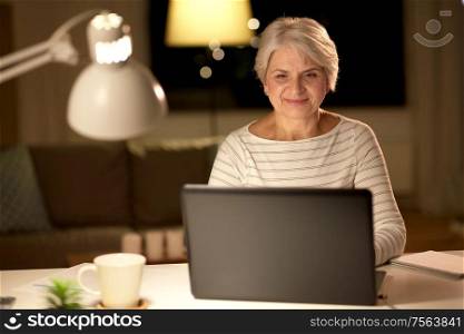 technology, old age and people concept - happy senior woman with laptop at home in evening. happy senior woman with laptop at home in evening