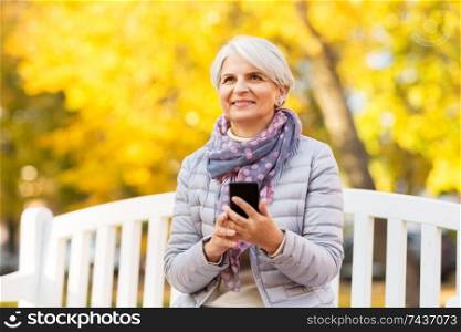 technology, old age and people concept - happy senior woman with smartphone sitting on bench at autumn park. happy senior woman with smartphone at autumn park