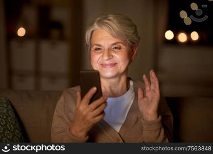 technology, old age and people concept - happy senior woman with smartphone having video call and waving hand at home in evening. happy old woman with smartphone having video call