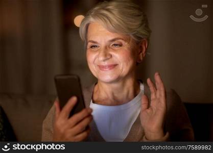 technology, old age and people concept - happy senior woman with smartphone having video call and waving hand at home in evening. happy old woman with smartphone having video call