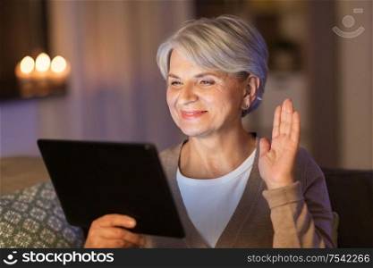 technology, old age and people concept - happy senior woman with tablet pc computer having video call and waving hand at home in evening. happy old woman with tablet pc having video call