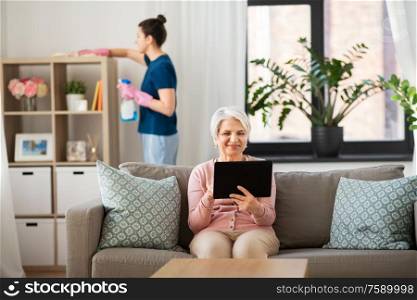 technology, old age and people concept - happy senior woman with tablet pc computer and housekeeper cleaning at home. old woman with tablet pc and housekeeper at home