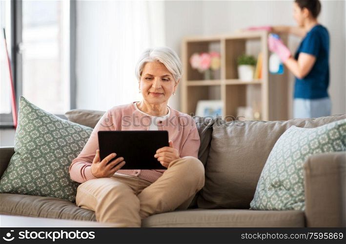 technology, old age and people concept - happy senior woman with tablet pc computer and housekeeper cleaning at home. old woman with tablet pc and housekeeper at home