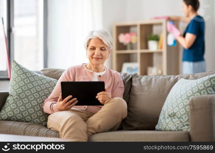 technology, old age and people concept - happy senior woman with tablet pc computer and housekeeper cleaning at home. old woman with tablet pc and housekeeper at home