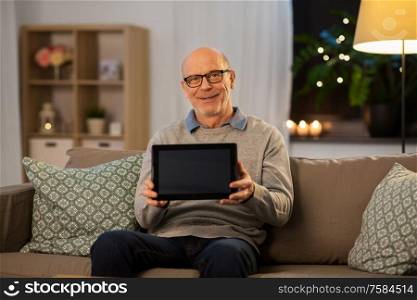 technology, old age and people concept - happy smiling bald senior man with tablet pc computer sitting on sofa at home in evening. happy senior man with tablet computer at home