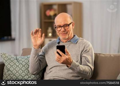 technology, old age and people concept - happy smiling bald senior man with smartphone having video call and waving hand at home in evening. old man with smartphone having video call at home