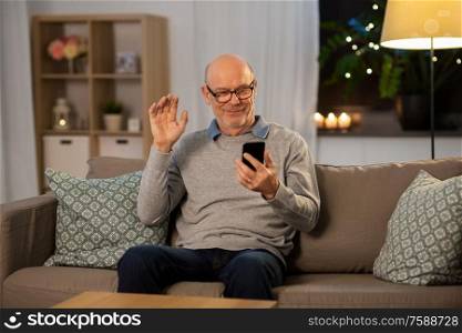 technology, old age and people concept - happy smiling bald senior man with smartphone having video call and waving hand at home in evening. old man with smartphone having video call at home