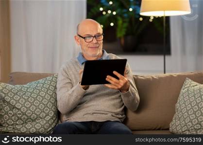 technology, old age and people concept - happy smiling bald senior man with tablet pc computer sitting on sofa at home in evening. happy senior man with tablet computer at home
