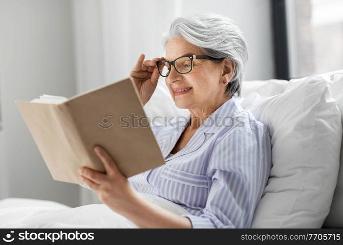 technology, old age and people concept - happy smiling senior woman in glasses reading book in bed at home bedroom. old woman in glasses reading book in bed at home