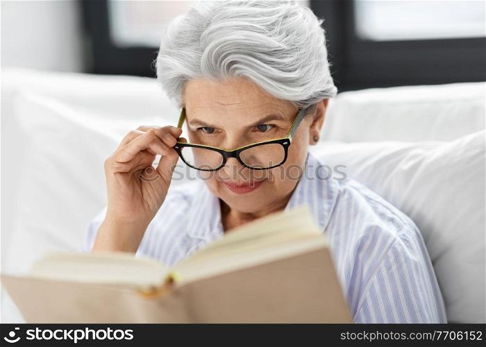 technology, old age and people concept - senior woman in glasses reading book in bed at home bedroom. old woman in glasses reading book in bed at home