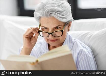 technology, old age and people concept - senior woman in glasses reading book in bed at home bedroom. old woman in glasses reading book in bed at home