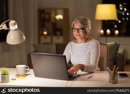 technology, old age and people concept - senior woman in glasses with laptop working at home in evening. senior woman with laptop working at home at night