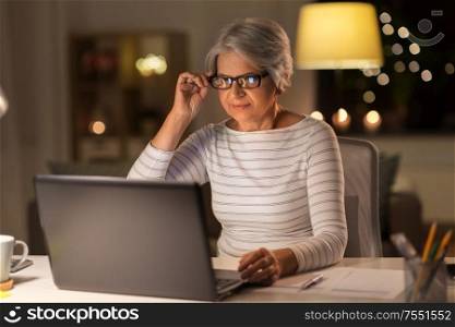 technology, old age and people concept - senior woman in glasses with laptop working at home in evening. senior woman with laptop working at home at night
