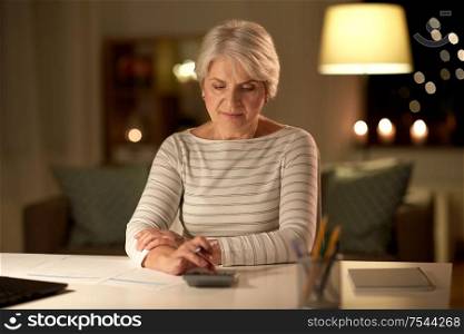 technology, old age and people concept - senior woman with laptop counting on calculator and filling tax form at home in evening. old woman counting on calculator at home at night