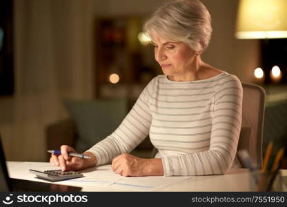 technology, old age and people concept - senior woman with laptop counting on calculator and filling tax form at home in evening. old woman counting on calculator at home at night