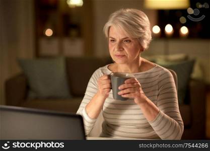 technology, old age and people concept - senior woman with laptop drinking coffee at home in evening. senior woman with laptop drinking coffee at home