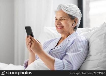 technology, old age and people concept - senior woman with smartphone and wireless earphones in bed at home bedroom. senior woman with smartphone and earphones in bed