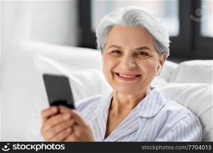technology, old age and people concept - senior woman with smartphone and wireless earphones in bed at home bedroom. senior woman with smartphone and earphones in bed